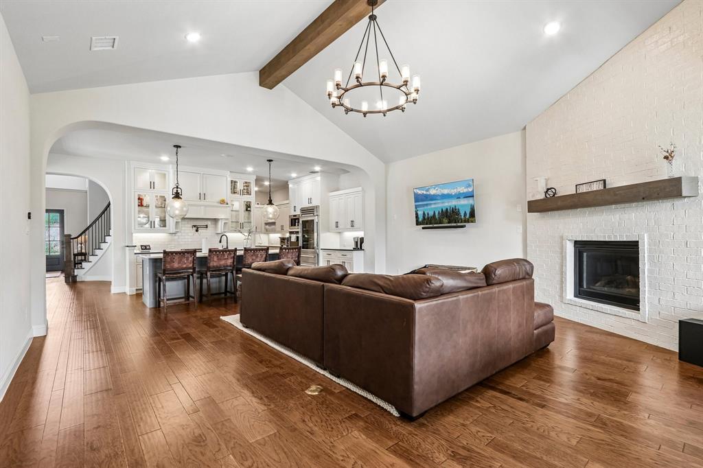 Vaulted ceiling with wood beam, wood-finish flooring, white brick fireplace with mantel, white kitchen cabinetry, and a kitchen island with pendant lighting