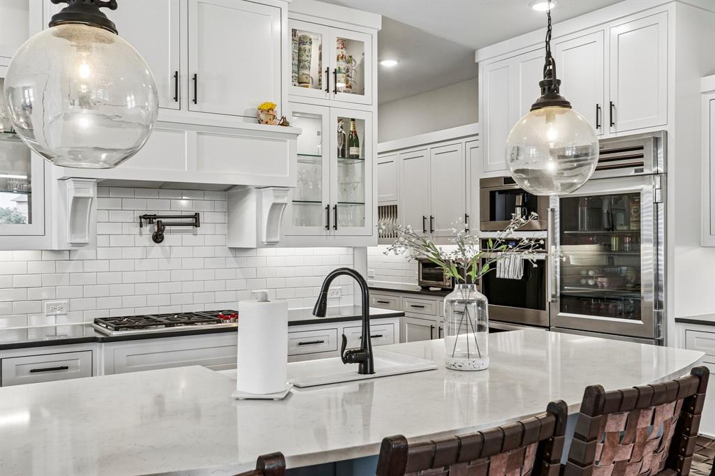 White shaker-style cabinetry with glass-front accents, subway tile backsplash, built-in stainless steel appliances, and a kitchen island with an undermount sink White shaker-style cabinetry with glass-front accents, subway tile backsplash, built-in stainless steel appliances, and a kitchen island with an undermount sink