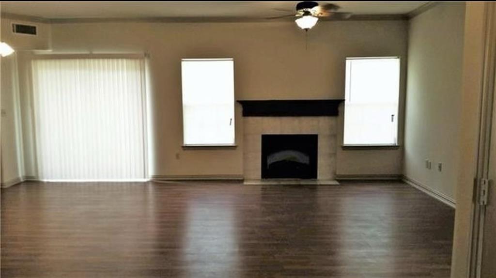 Spacious room featuring wood-finish flooring, a fireplace with a stone surround and dark mantel, a ceiling fan with integrated lighting, and multiple windows with vertical blinds