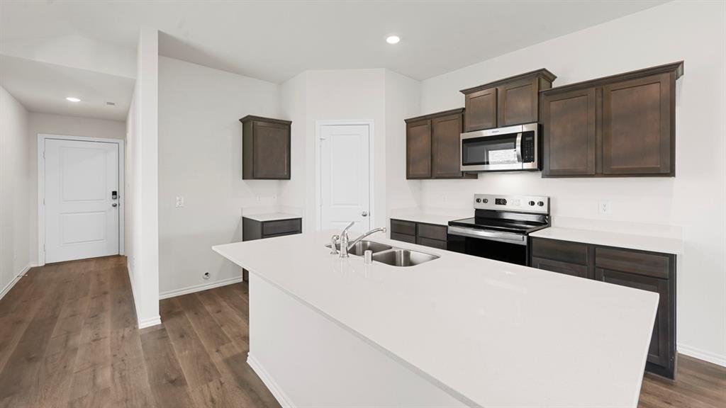 Kitchen featuring a large island with a double basin sink, dark wood-finish cabinetry, light-toned countertops, stainless steel appliances, and wood-finish flooring Kitchen featuring a large island with a double basin sink, dark wood-finish cabinetry, light-toned countertops, stainless steel appliances, and wood-finish flooring