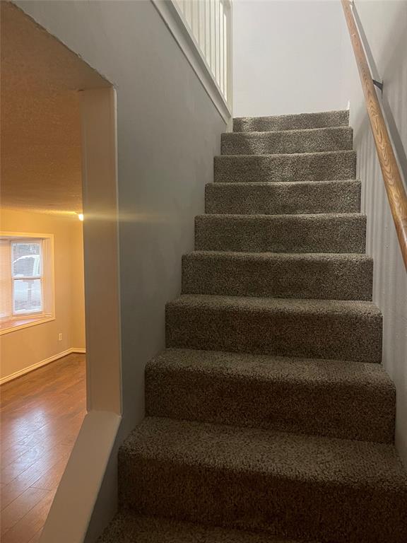 Carpeted staircase featuring a wooden handrail and white vertical balusters, accompanied by wood-finish flooring and a bay window in the foreground