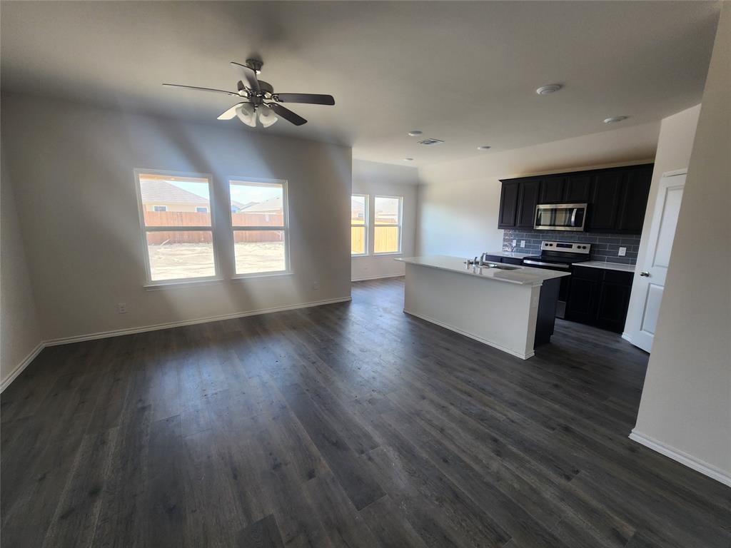 Spacious open-concept layout featuring wood-finish flooring, a kitchen island with a built-in sink, dark cabinetry, and a subway tile backsplash