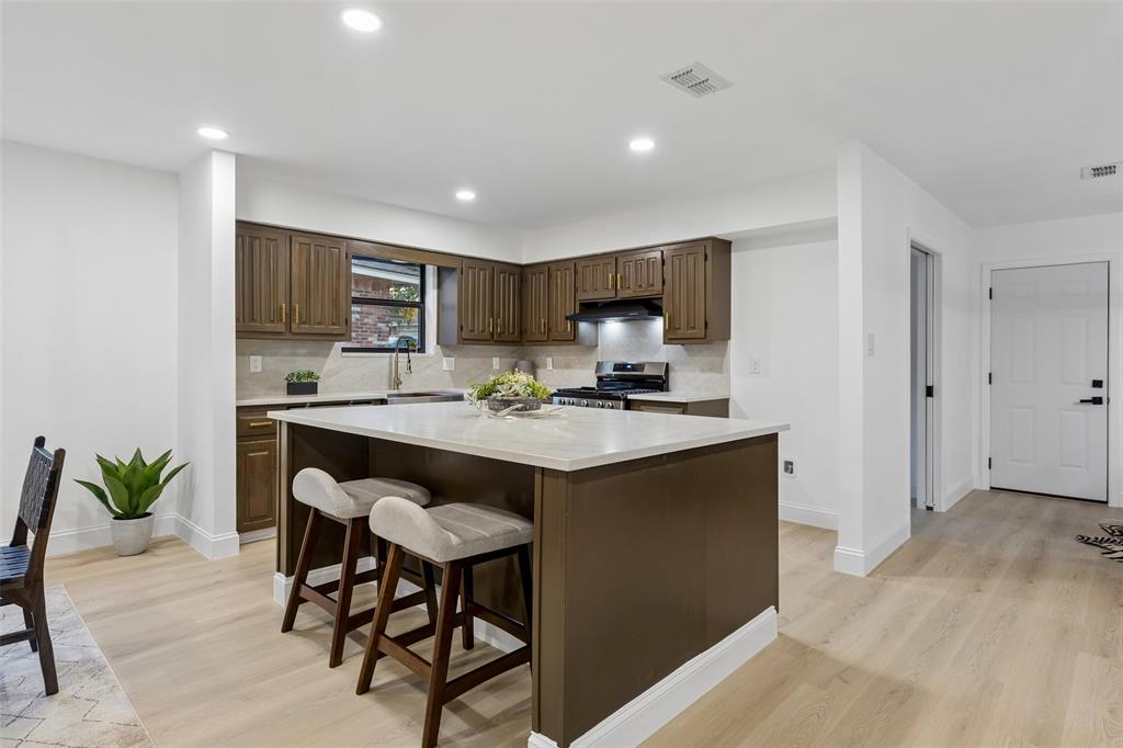Central kitchen island with a light-toned countertop, wood-finish flooring, recessed lighting, brown cabinetry, and a tile backsplash