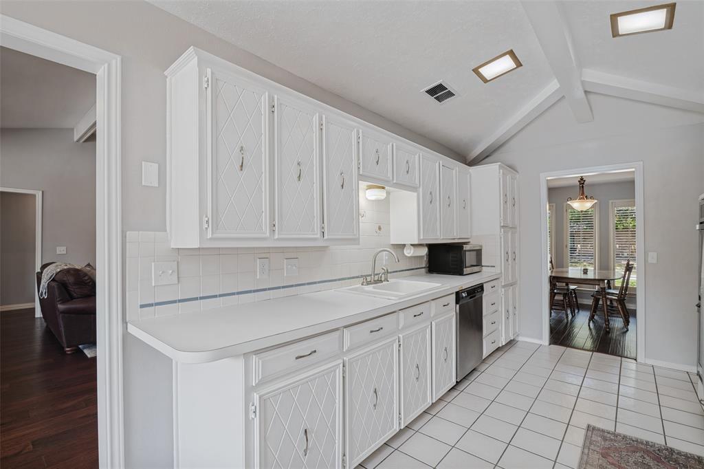 White cabinetry with diamond pattern detailing, white tile backsplash, vaulted ceiling with exposed beams, stainless steel dishwasher, and tile flooring