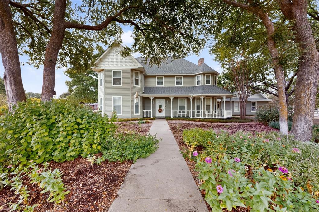 Two-story exterior with light green siding and white trim, featuring a wrap-around porch with white railings, a prominent gabled dormer, and established landscaping with mature trees and flowering plants Two-story exterior with light green siding and white trim, featuring a wrap-around porch with white railings, a prominent gabled dormer, and established landscaping with mature trees and flowering plants