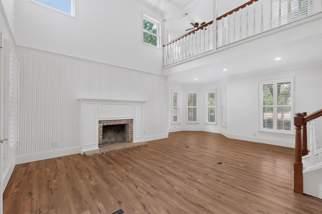 Two-story living area with wood-finish flooring, a brick fireplace with a white mantel, and a bay window featuring plantation shutters