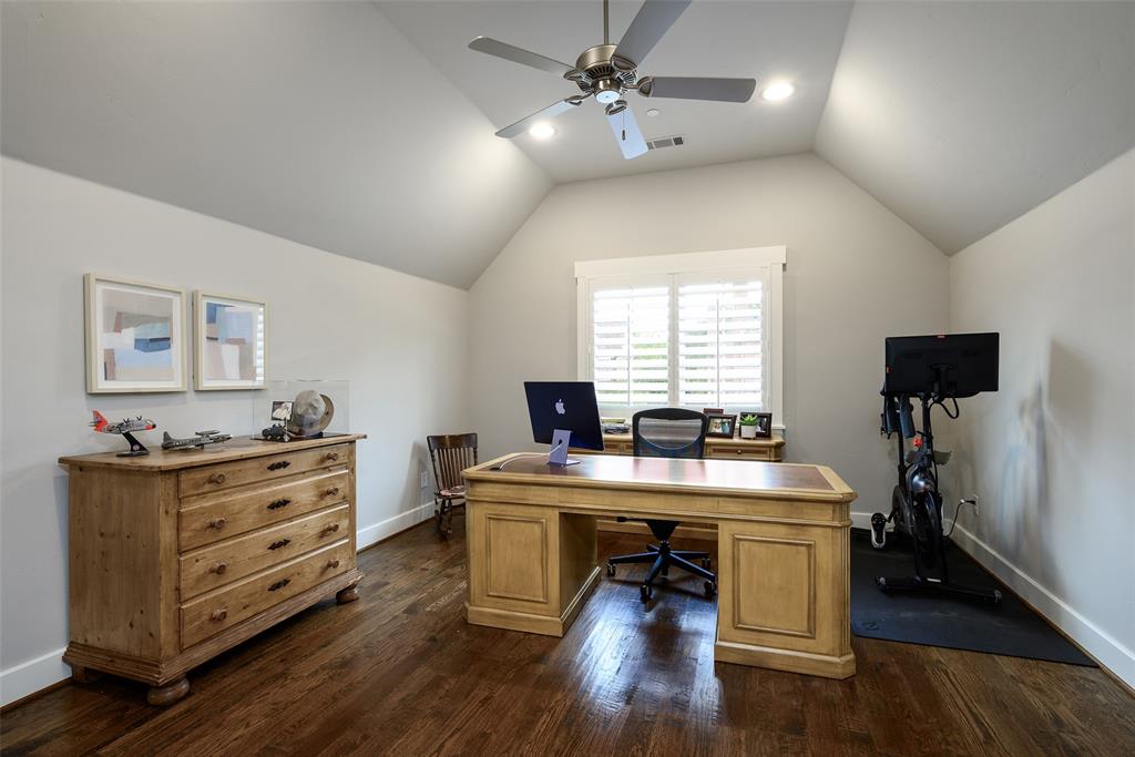 Spacious home office featuring vaulted ceilings with ceiling fan and recessed lighting, hardwood flooring, dual wooden desks with ample storage cabinetry, and shuttered windows flooding the room with natural light