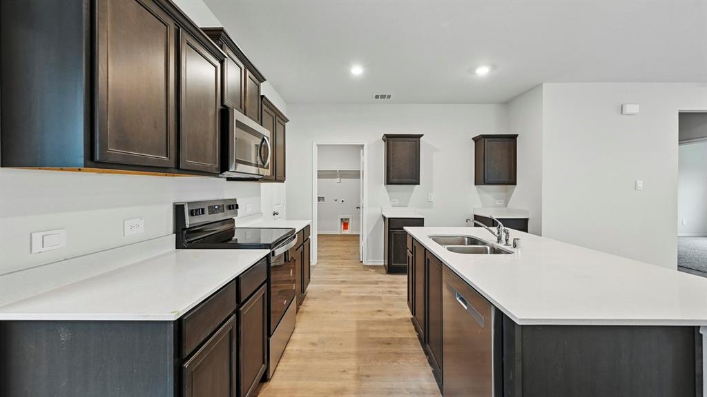 Open-concept kitchen featuring an expansive island with a double basin sink, dark wood-finish cabinetry, stainless steel appliances, light countertops, and wood-finish flooring