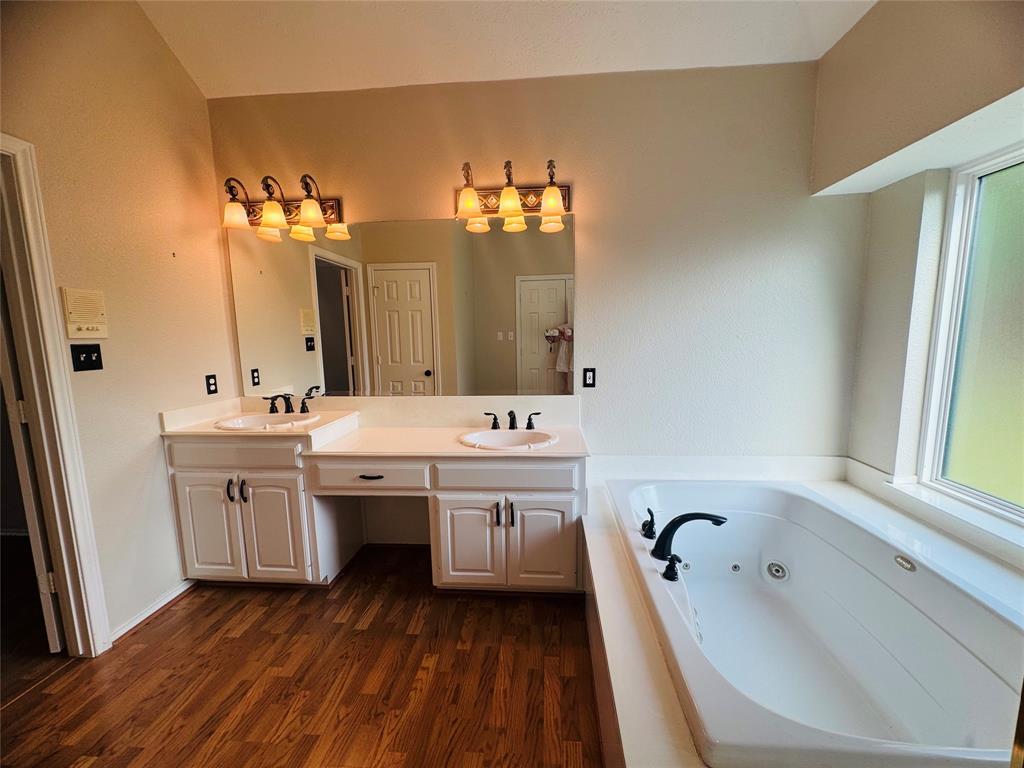 Spa-inspired primary bathroom showcasing dual vanities with ample storage cabinetry, illuminated mirror sconces, soaking tub with deck surround positioned at the window, wood-finish flooring, and matte black faucet hardware throughout