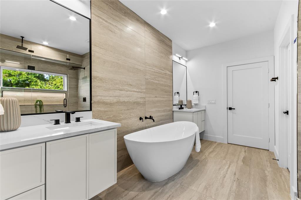 Contemporary bathroom featuring a freestanding soaking tub, dual vanities with white cabinetry and integrated sinks, a walk-in shower with a rain shower head, and floor-to-ceiling textured tile accents