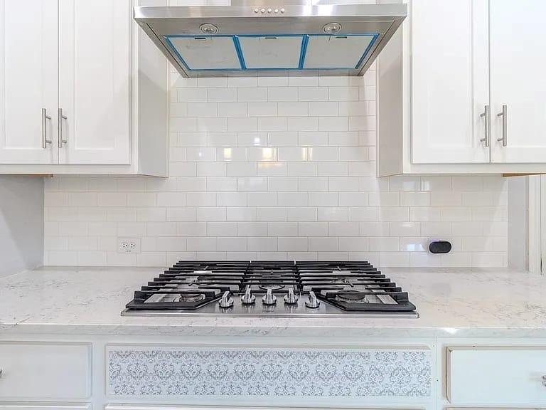 Stainless steel gas range with a coordinating hood, white subway tile backsplash, white shaker-style cabinetry, and light-toned countertops