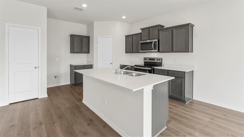 Bright kitchen featuring a central island with an integrated sink, dark gray cabinetry, stainless steel appliances, and wood-finish flooring