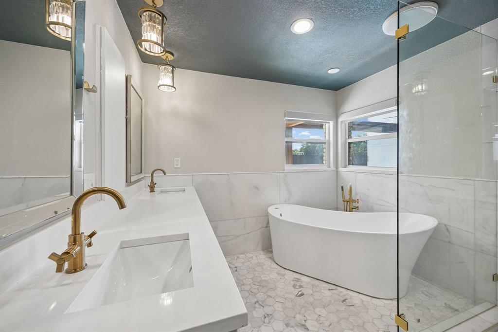 Beautifully updated primary bath featuring marble finishes, dual vanities, a freestanding soaking tub, glass-enclosed shower and elegant gold fixtures.