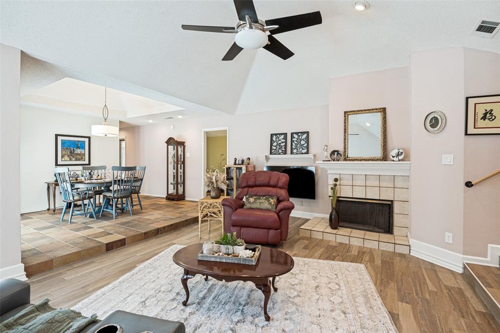 Vaulted ceiling with a ceiling fan, tiled fireplace with a mantel, wood-finish flooring, and a dining area featuring a tray ceiling and a pendant light fixture