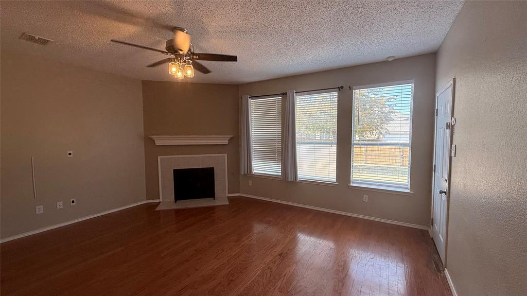 Spacious room featuring wood-finish flooring, a ceiling fan with integrated lighting, and a corner fireplace with a white mantel and tile surround