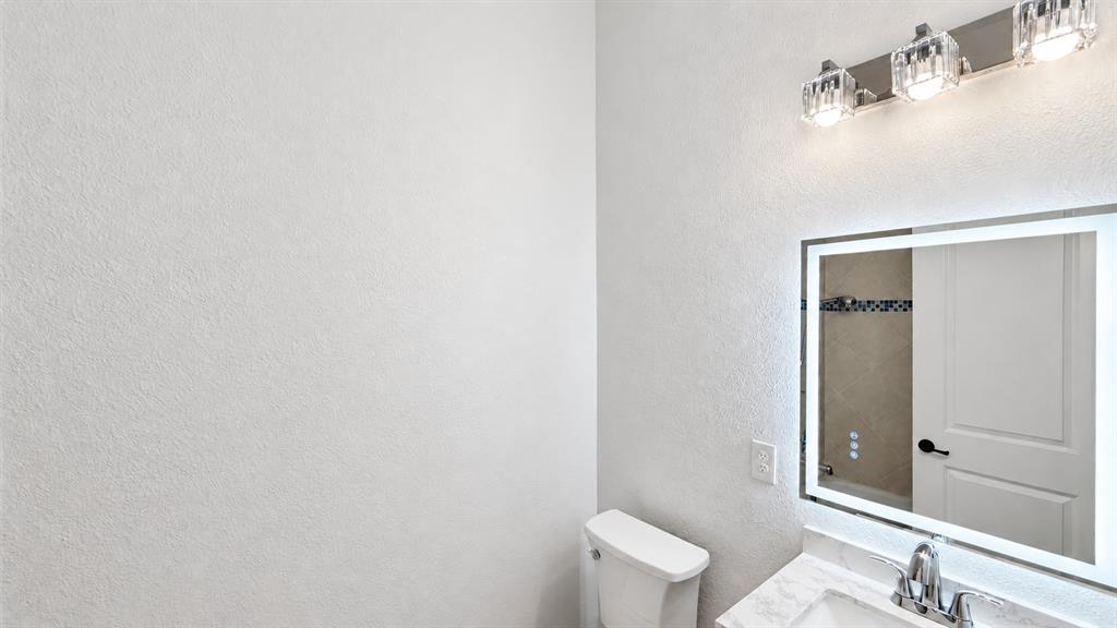 Backlit vanity mirror, modern chrome faucet, white toilet, crystal-accented light fixture, and textured wall finish