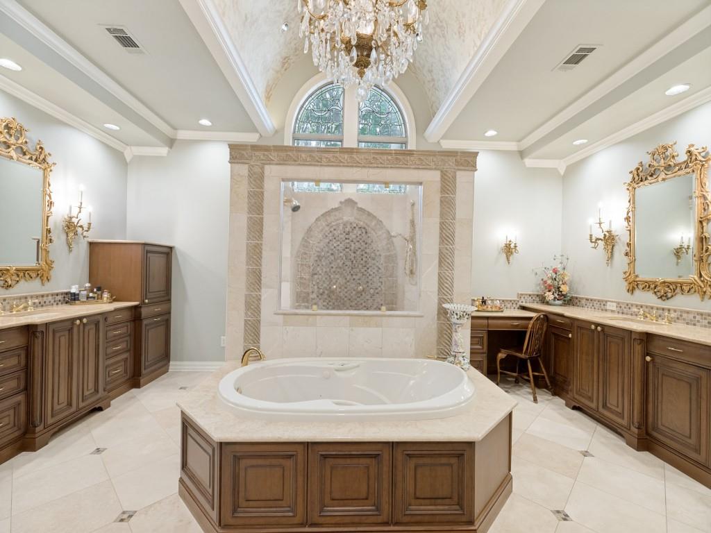 Vaulted ceiling with an ornate chandelier, a built-in jetted tub with stone surround, dual vanities with wood-finish cabinetry, and a walk-in shower with mosaic tile