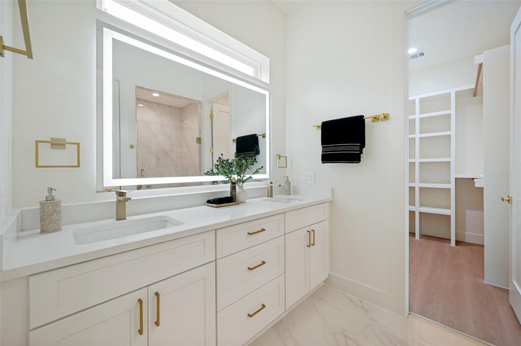 Dual vanity with white cabinetry and gold-finish hardware, illuminated mirror, and a walk-in closet with built-in shelving