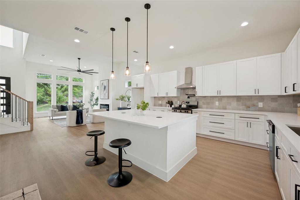 Expansive open concept living space featuring wood-finish flooring, a contemporary kitchen with a large island, stainless steel appliances, and white cabinetry, complemented by a living area with a fireplace and large windows