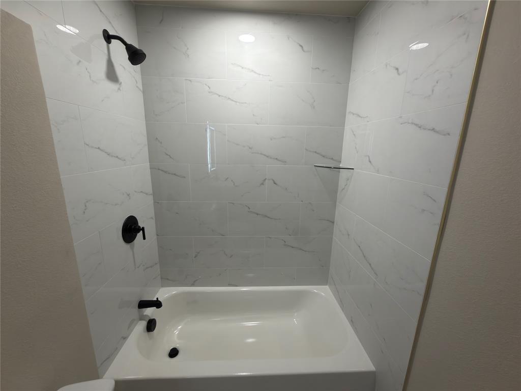 Bathtub with white tiled surround, matte black shower fixtures, and recessed glass shelf