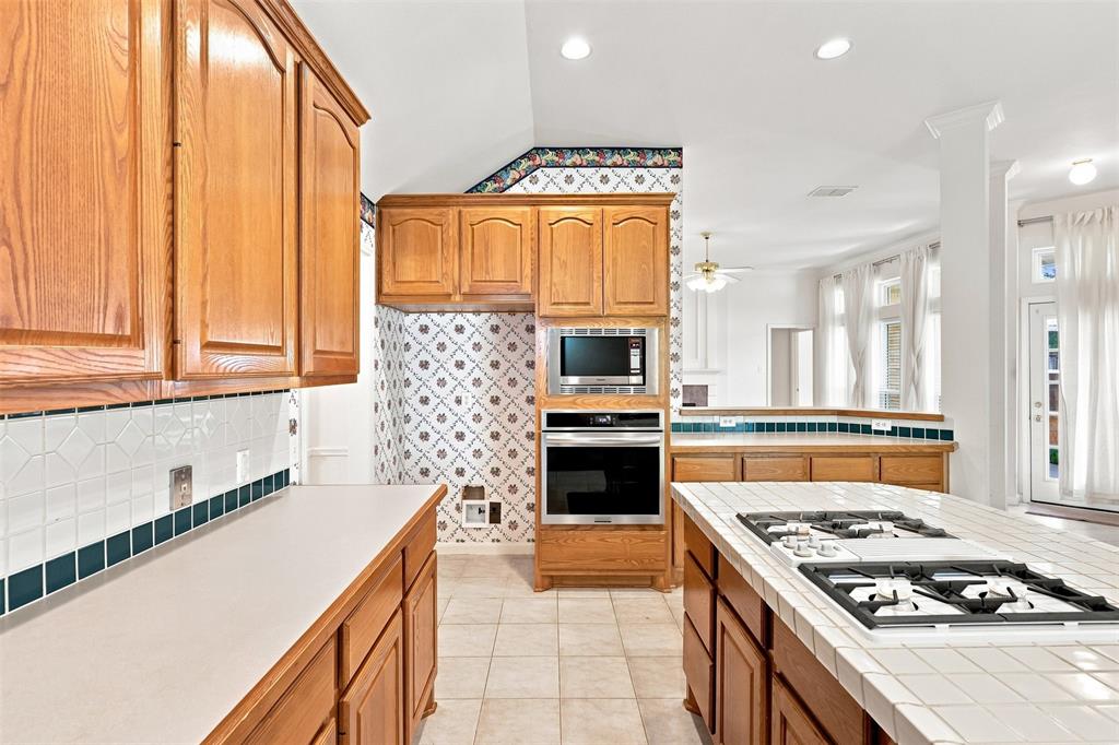 Wood cabinetry with stainless steel built-in appliances, tile backsplash, and a tile-top island with a cooktop.