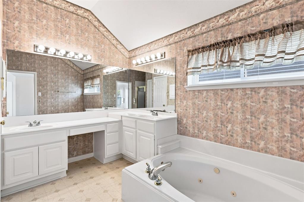 Primary bathroom features dual vanity sinks with white cabinetry, mirrored wall, recessed jetted bathtub, and patterned wallpaper.