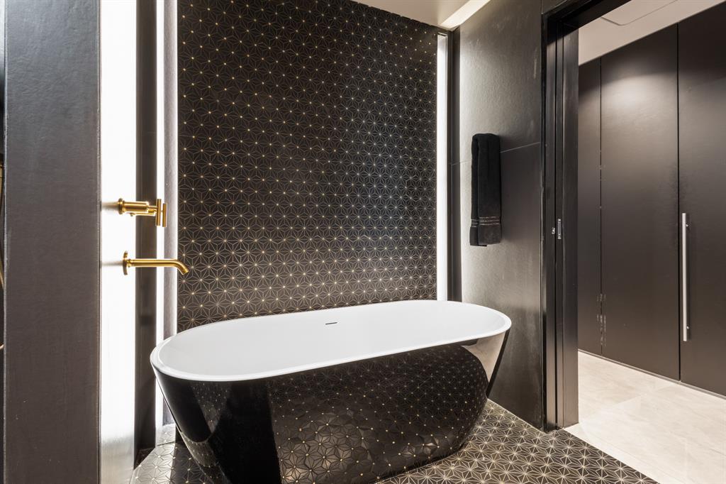 Freestanding black and white soaking tub, geometric pattern wall and floor tiles, gold-finish wall-mounted faucet, integrated vertical lighting, and built-in matte black cabinetry