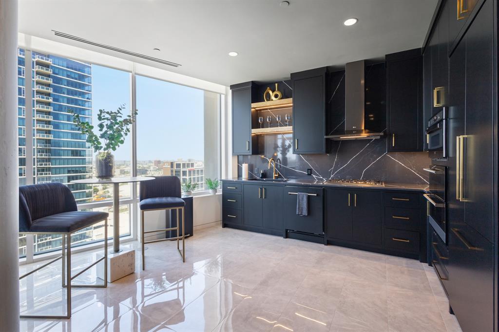 Sleek kitchen featuring dark cabinetry, integrated appliances, gold-finish hardware, a full-height backsplash with veining, and a gas cooktop Sleek kitchen featuring dark cabinetry, integrated appliances, gold-finish hardware, a full-height backsplash with veining, and a gas cooktop