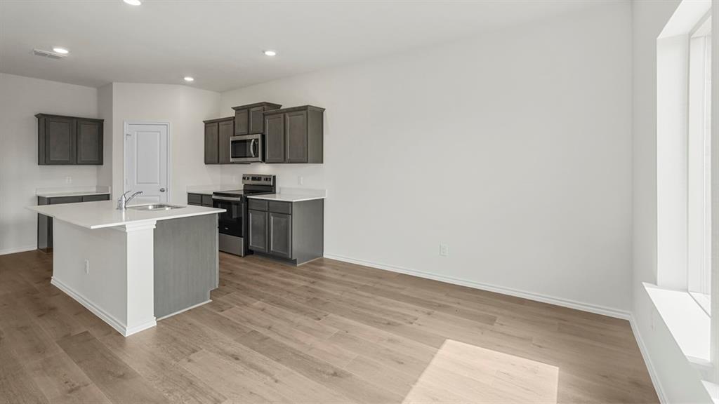 Open-concept kitchen featuring dark gray cabinetry, white countertops, stainless steel appliances, a central island with an undermount sink, and wood-finish flooring