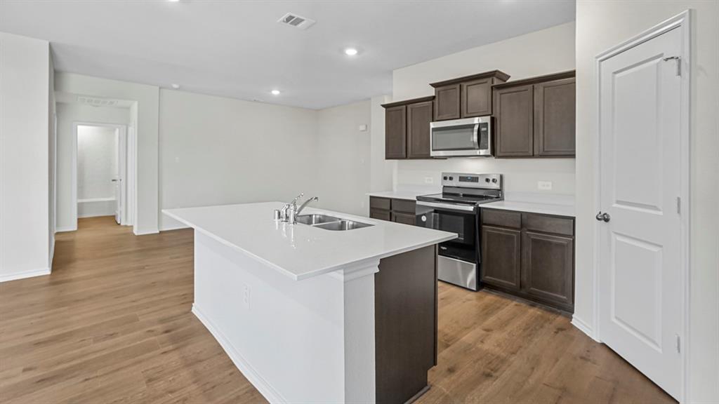 Kitchen island with dual basin sink, integrated stainless steel appliances, dark wood cabinetry, and wood-finish flooring
