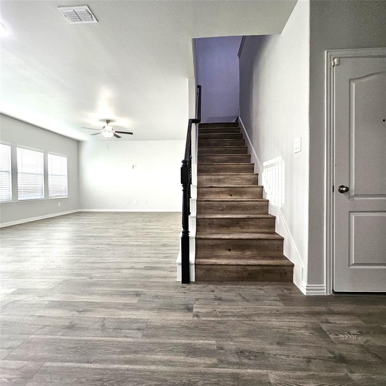 Spacious living area featuring wood-finish flooring, multiple windows with blinds, a ceiling fan, and a staircase with a dark wood-finish handrail