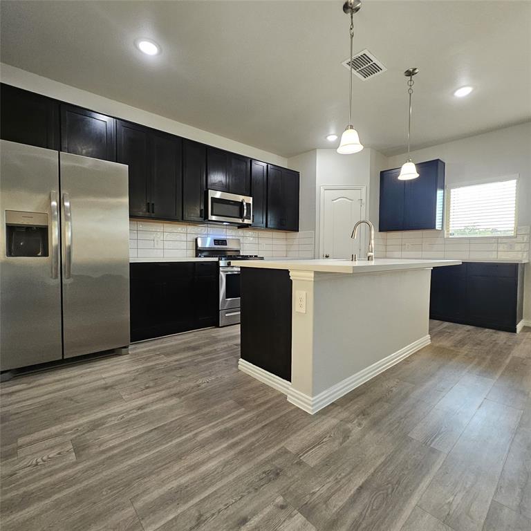 Kitchen with wood-finish flooring, dark cabinetry, stainless steel appliances, a white subway tile backsplash, and an island with a built-in sink
