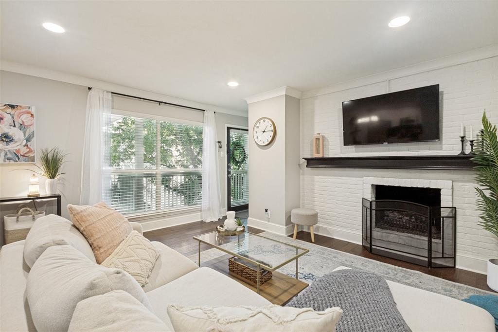 Recessed lighting, wood-finish flooring, white brick fireplace with a dark mantel, large window with blinds, and crown molding