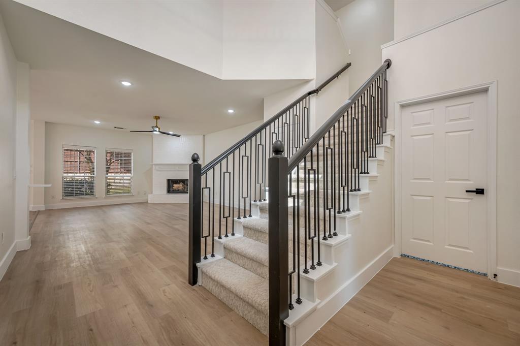 Luxury Vinyl Plank flooring, staircase with wrought iron balusters, carpeted stair treads, ceiling fan with integrated lighting, and a fireplace with a white mantel