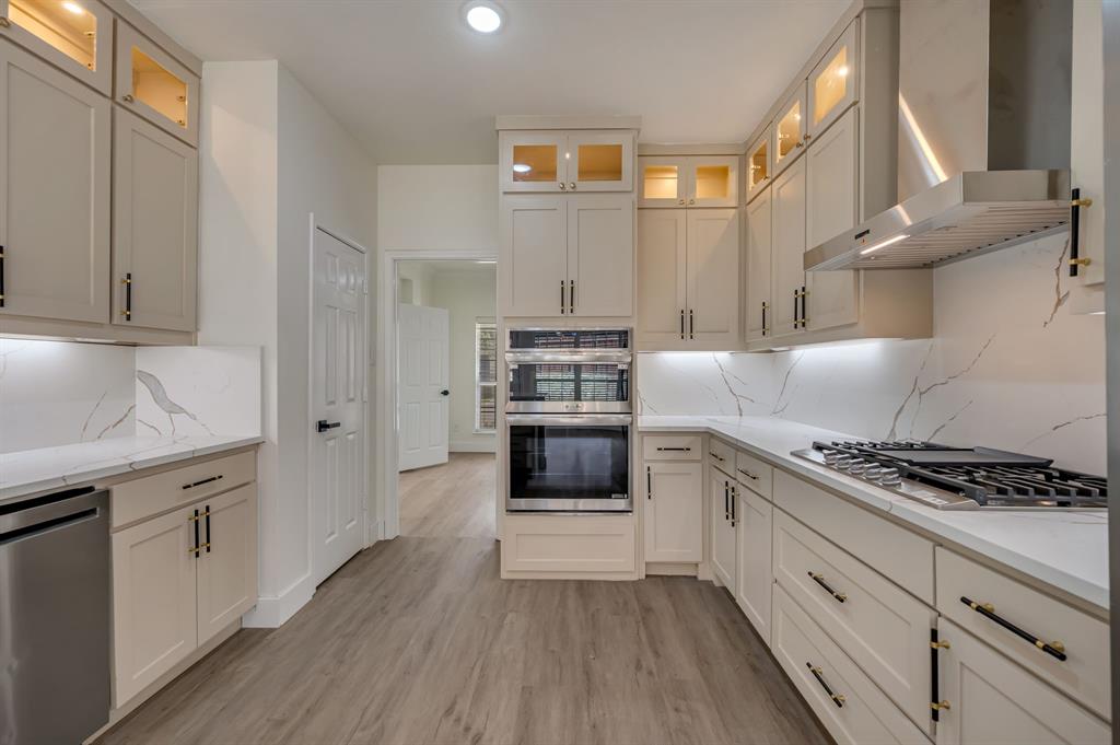 Gourmet kitchen featuring a built-in double oven, gas cooktop, stainless steel range hood, stone countertops with matching backsplash, and custom cabinetry with glass-front uppers Gourmet kitchen featuring a built-in double oven, gas cooktop, stainless steel range hood, stone countertops with matching backsplash, and custom cabinetry with glass-front uppers
