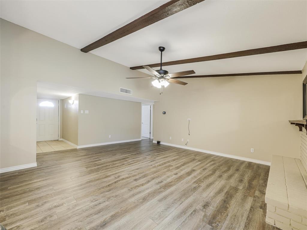 Vaulted ceiling with exposed wood beams, wood-finish flooring, ceiling fan with integrated lighting, brick fireplace with mantel, and recessed entry
