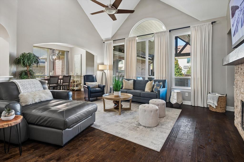 Vaulted ceiling with a ceiling fan, dark wood-finish flooring, stone-clad fireplace with a wood mantel, and tall windows