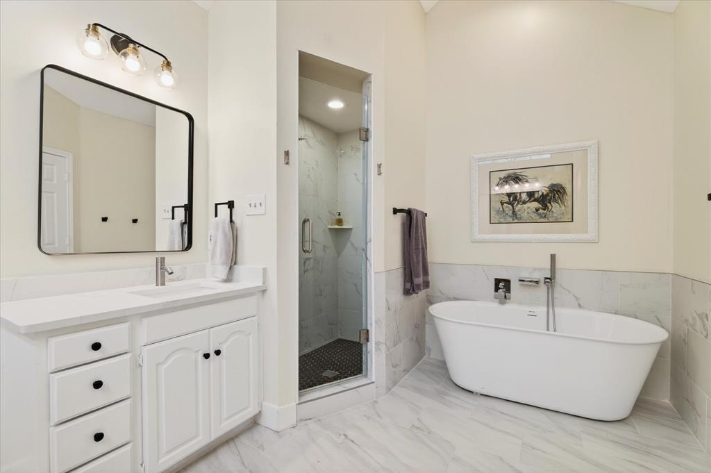 White vanity with a solid surface countertop, separate glass-enclosed shower with tiled walls, freestanding soaking tub, and large format floor tiling