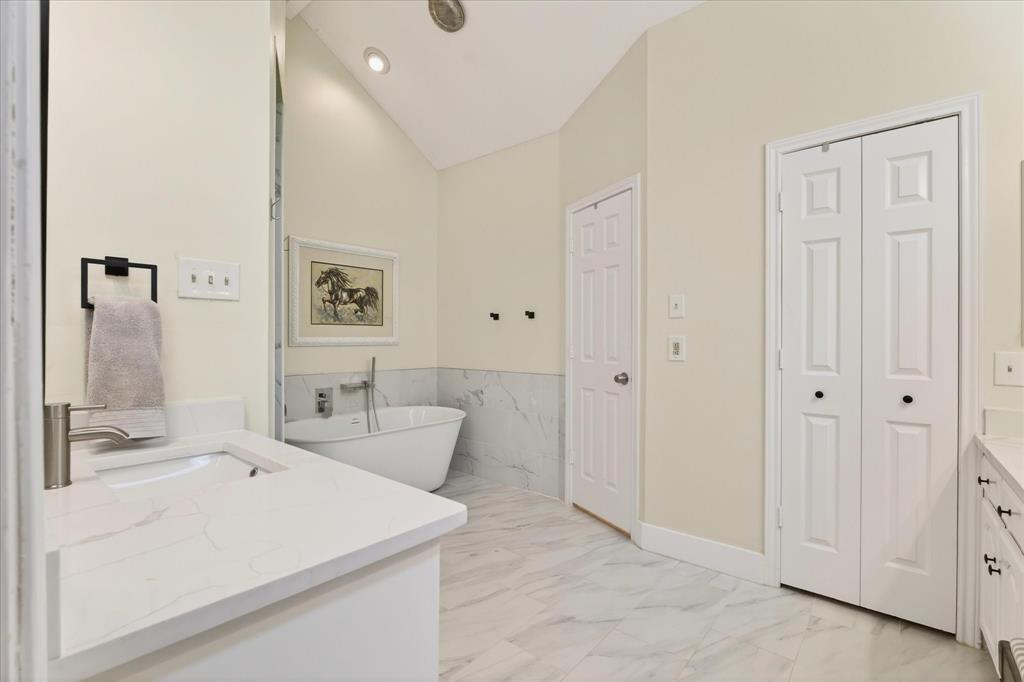 Freestanding soaking tub with wall-mounted faucet, marble-look tiled floors, white vanity with integrated sink and stone-look countertop, and recessed lighting