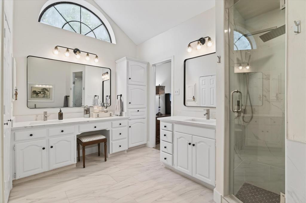 Vaulted ceiling with an arched window, dual vanities with white cabinetry and solid surface countertops, built-in storage cabinet, and a glass-enclosed shower