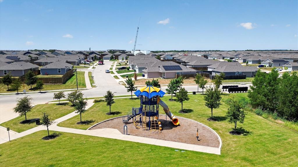 Community green space featuring a playground with slides and climbing elements, surrounded by a manicured lawn, paved walkways, and new construction homes