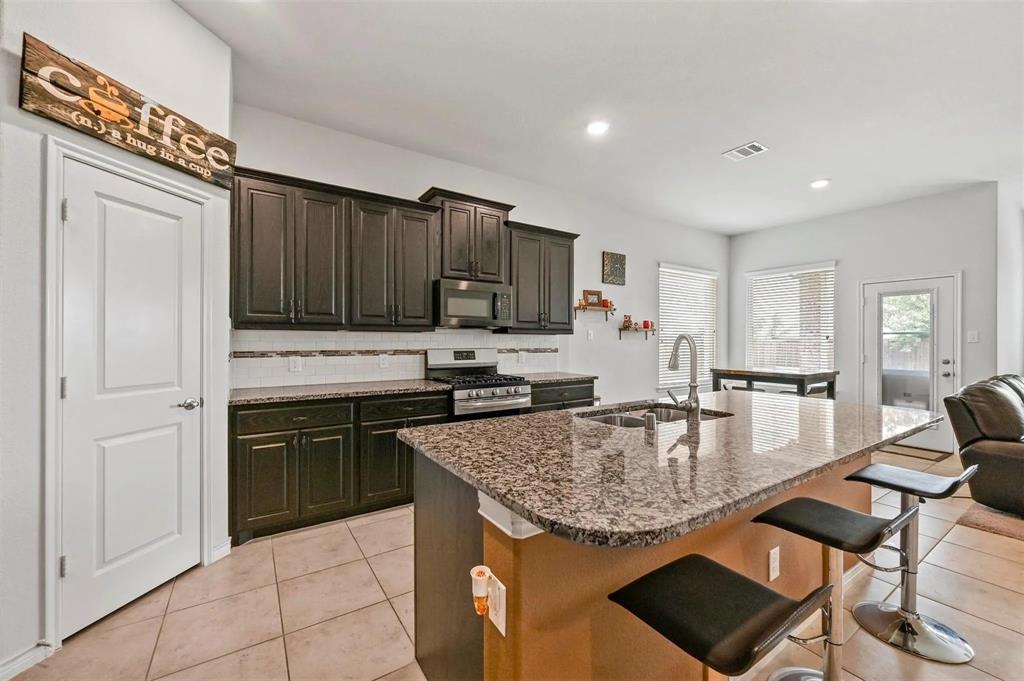 Kitchen featuring dark wood-finish cabinetry, stainless steel appliances, a multi-tone tile backsplash, and an island with a polished stone countertop and integrated sink Kitchen featuring dark wood-finish cabinetry, stainless steel appliances, a multi-tone tile backsplash, and an island with a polished stone countertop and integrated sink