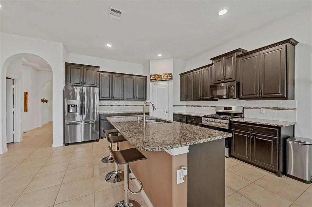 Kitchen featuring dark wood-finish cabinetry, stainless steel appliances, granite countertops, a large kitchen island with a sink, and tile flooring