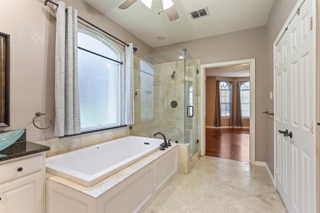 Bathroom featuring a drop-in bathtub with stone-finish surround, a glass-enclosed shower with tiled walls, an arched window, and stone-finish flooring