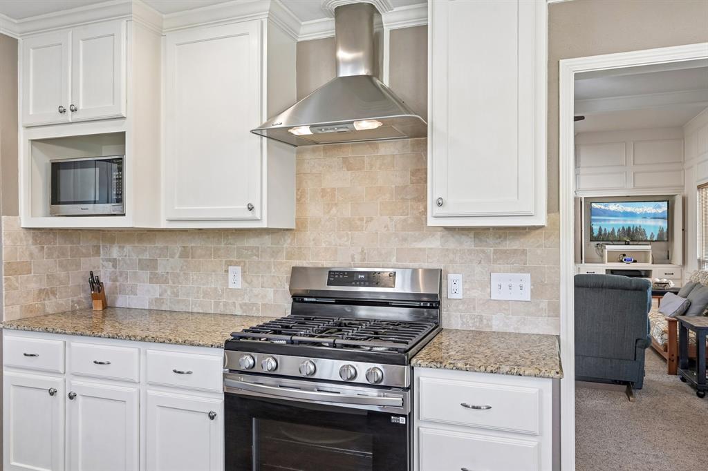 White cabinetry with crown molding, stainless steel range with overhead hood, integrated microwave, granite-finish countertops, and natural stone tile backsplash