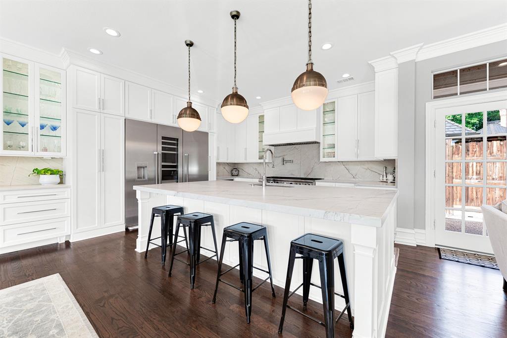 Crisp white cabinetry paired with quartzite countertops frames a oversized center island with breakfast bar seating. High-end stainless steel Thermador appliances, glass-front upper displays, and chic globe pendant lights complete this bright, contemporary kitchen workspac