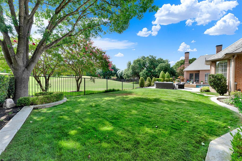 A manicured green lawn framed by wrought-iron fencing offers unobstructed views of the adjacent golf course fairway. Mature shade trees and stone-paved walkways border the yard, creating a tranquil transition between the private lawn and the rear patio entertaining area.