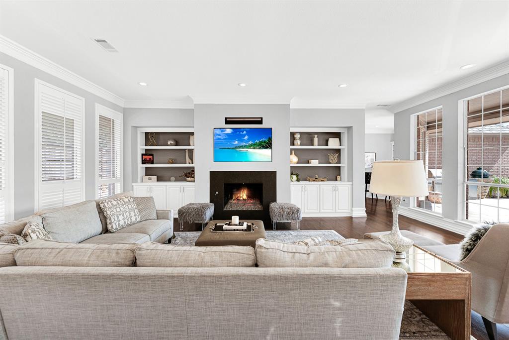 A sleek black fireplace hearth flanked by custom inset shelving units forms the visual anchor of this bright family space. Wall-to-wall windows paired with white plantation shutters flood the room with natural light while offering views of the covered patio.