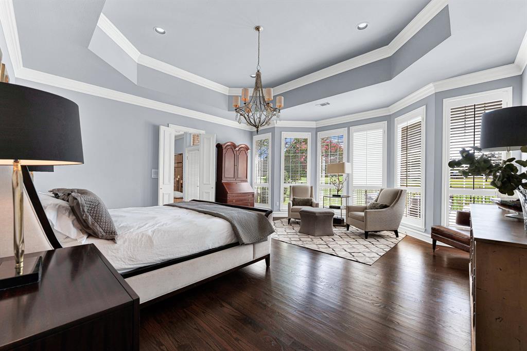Rich hardwood floors and an elegant tray ceiling with a rustic chandelier set a sophisticated tone in this expansive primary retreat. A sunlit bay window alcove with plantation shutters easily accommodates a private sitting area overlooking the manicured grounds.