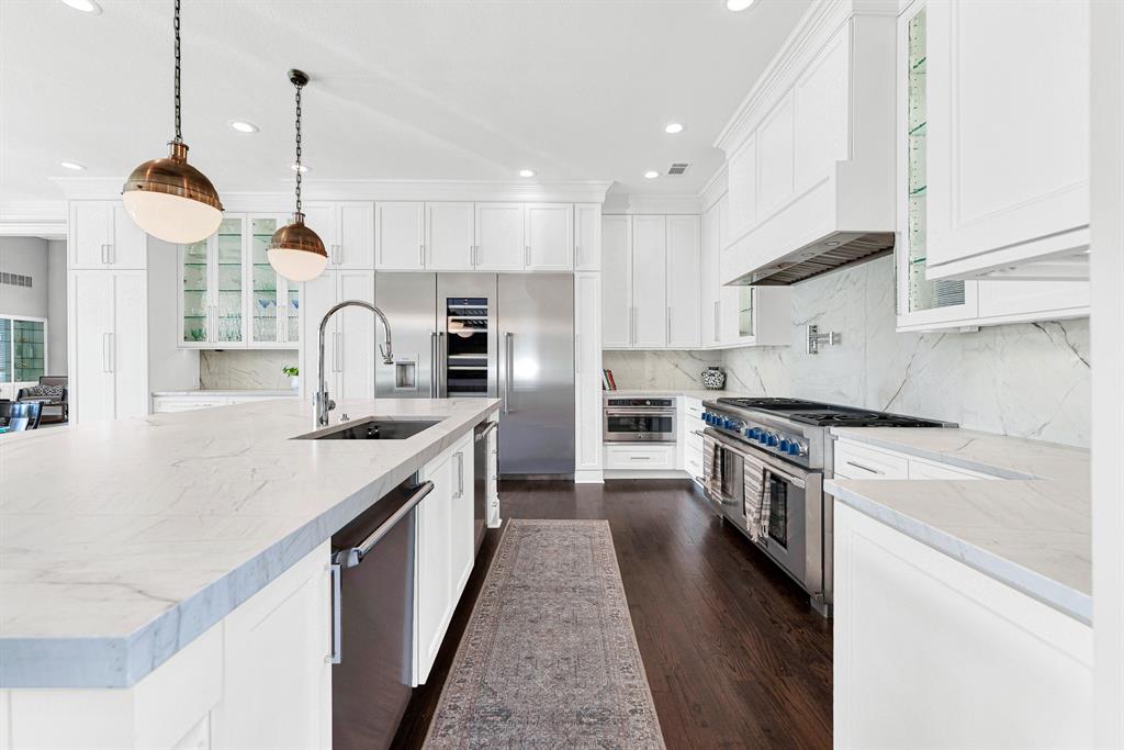 A commercial-grade gas range with double ovens and custom vent hood stand ready for meal prep alongside seamless slab backsplashes and built-in microwave. Thoughtful layout planning places the undermount sink and dual dishwashers directly within the expansive prep island.